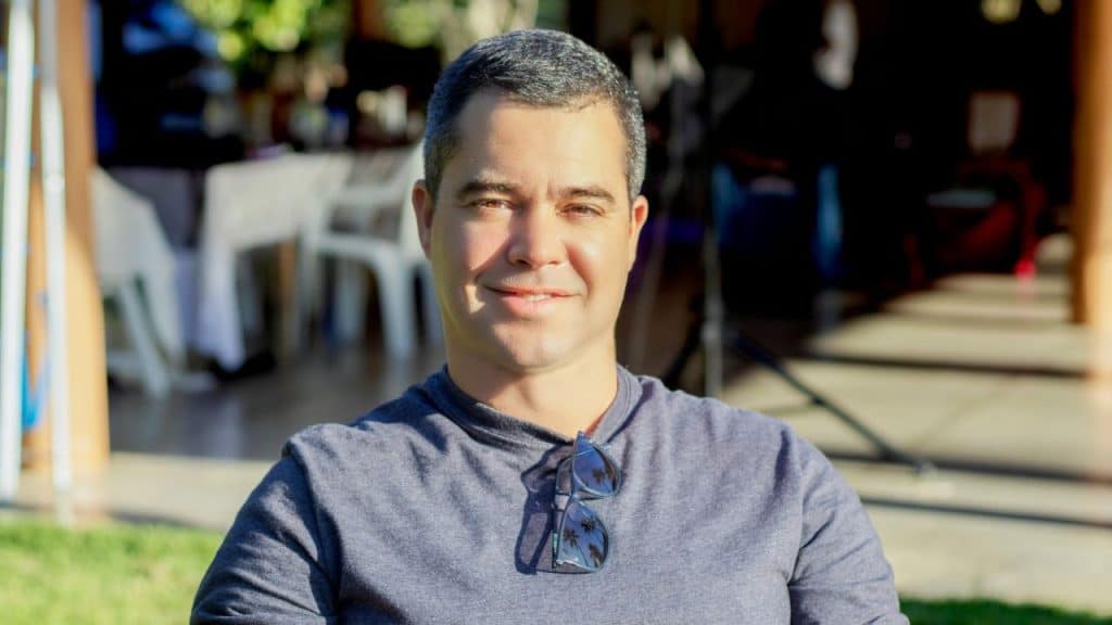 Smiling man with graying hair and sunglasses on his shirt sitting outdoors in the sun.