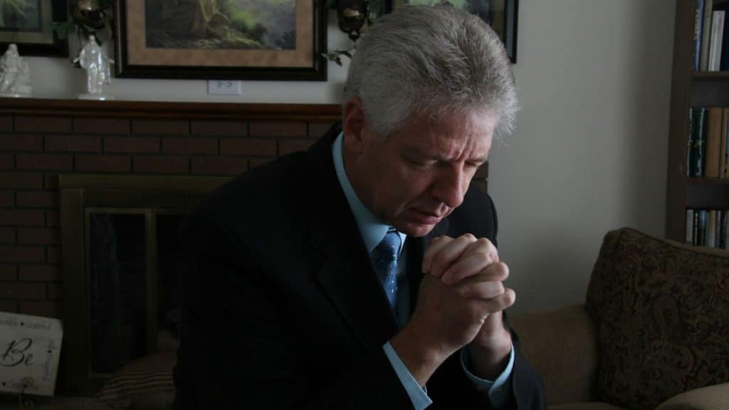 Older man in a suit with gray hair sits, clasping his hands and looking down.