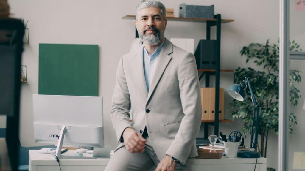 Well-dressed, gray-haired man with a beard sitting on a desk in a modern office, looking at the camera.