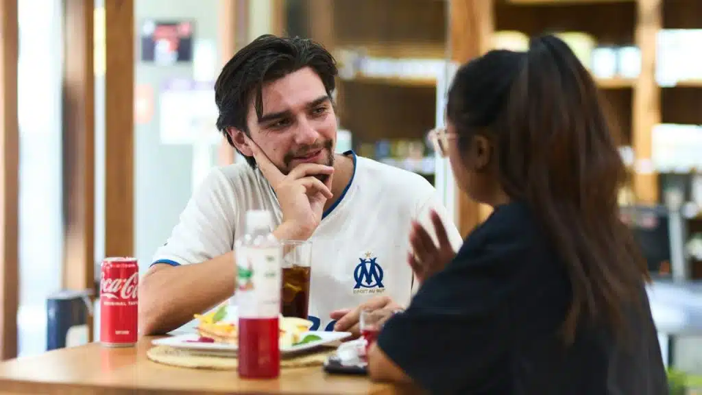A smiling man wearing a white and blue soccer jersey attentively listens to a woman at a table.