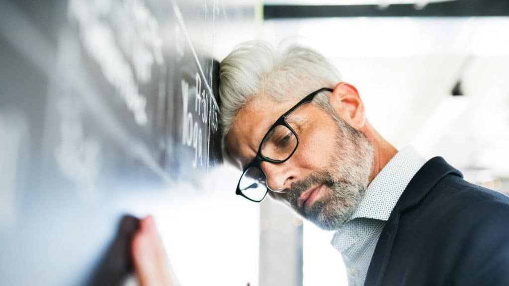 Stressed, middle-aged man with gray hair and glasses leaning his forehead against a dark wall or chalkboard.