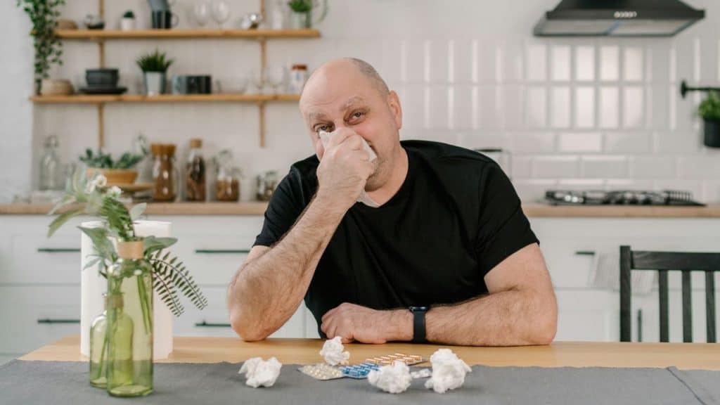 Man wiping his nose with a tissue at a table with crumpled tissues and medicine.