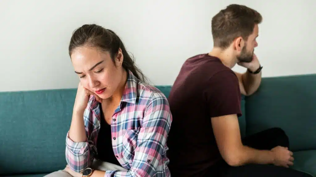Man and woman sitting back-to-back on a teal couch, each resting their head in a hand.