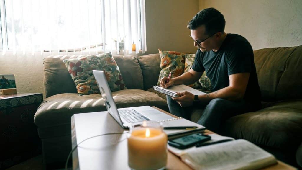 A man sits on a couch, writing in a notebook next to a laptop and a lit candle.
