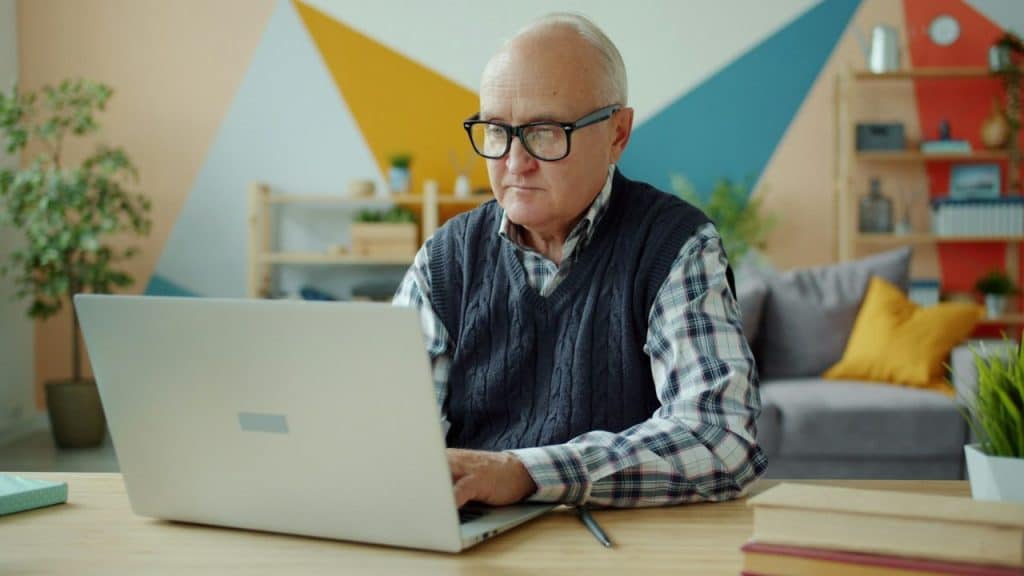 Older man in a plaid shirt and dark sweater vest wearing glasses, working intently on a laptop at a desk.
