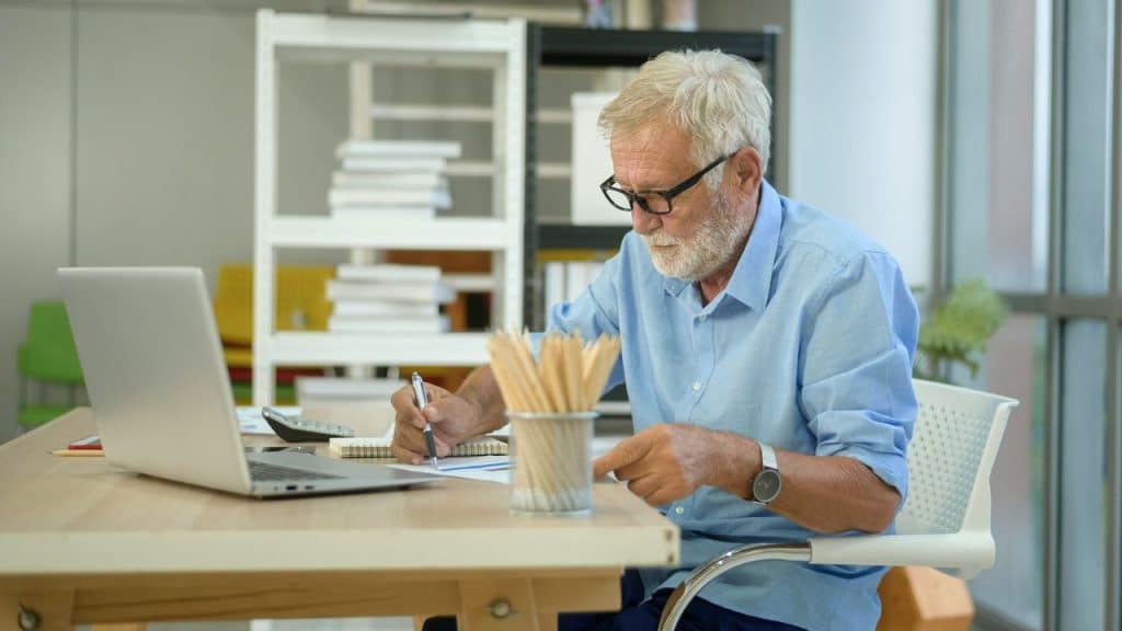 Elderly man with glasses writing notes at a wooden desk with a laptop.
