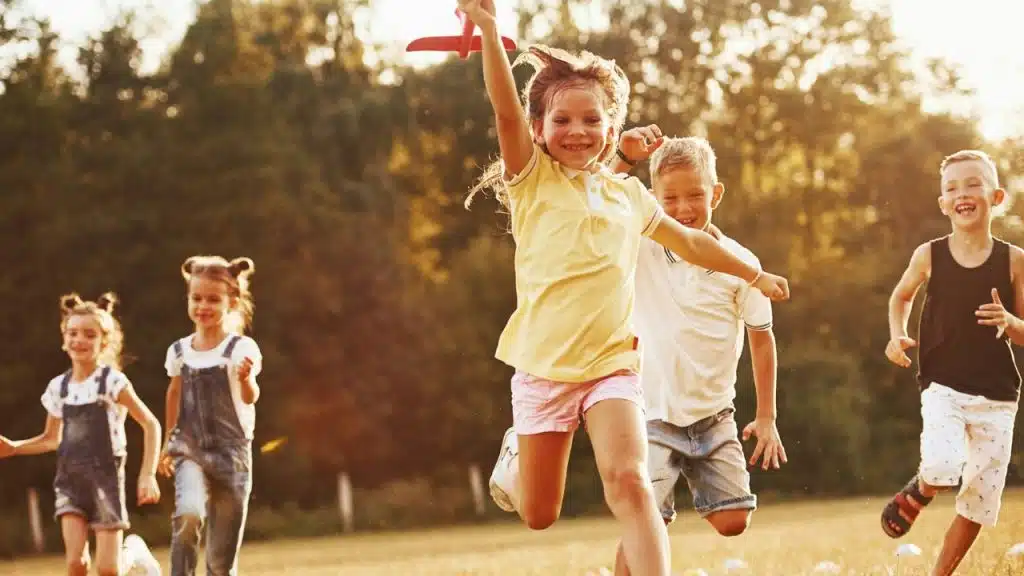 Five smiling children running through a sunny grassy field with trees in the background.
