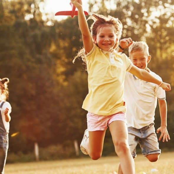 Five smiling children running through a sunny grassy field with trees in the background.