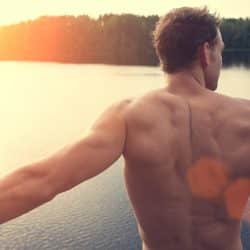 A muscular, shirtless man standing in front of a lake.