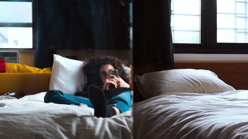 A woman wearing jeans and socks is relaxing in bed, looking at her smartphone.