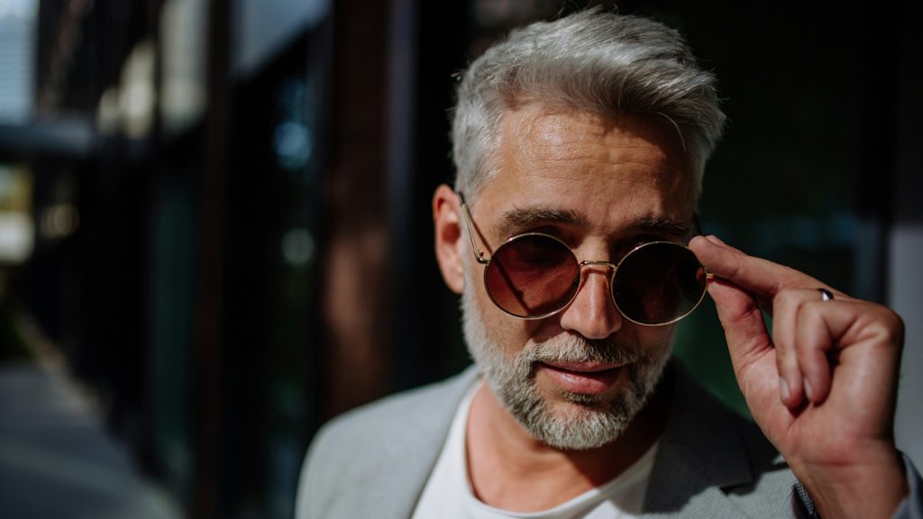 A bearded man in a grey suit adjusting his sunglasses.