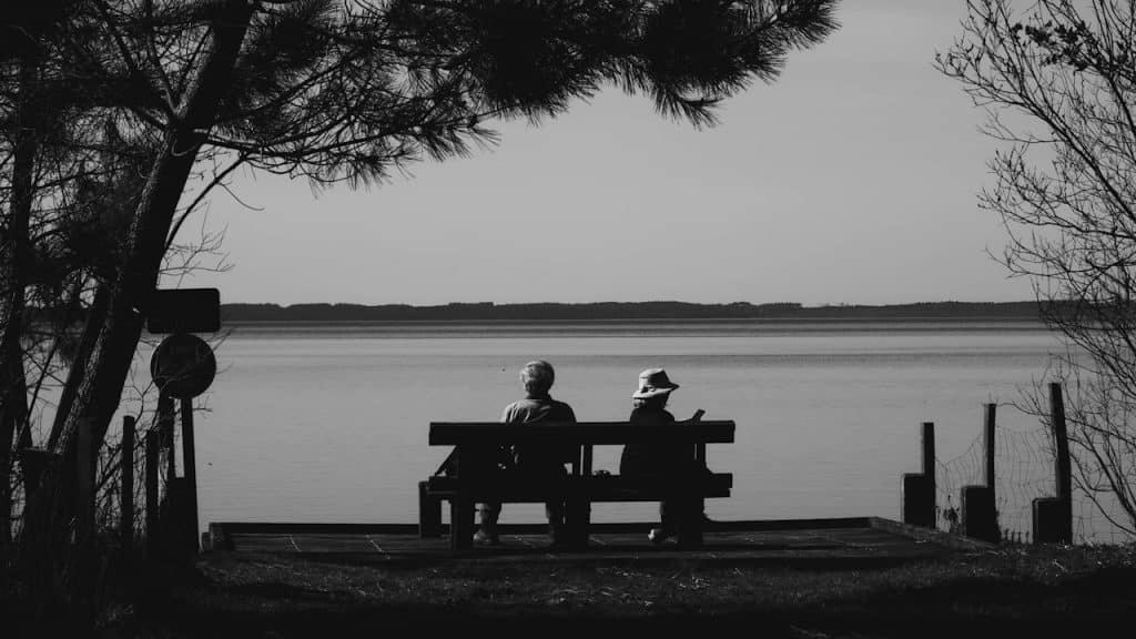 A couple sitting apart from each other on a bench beside a lake.