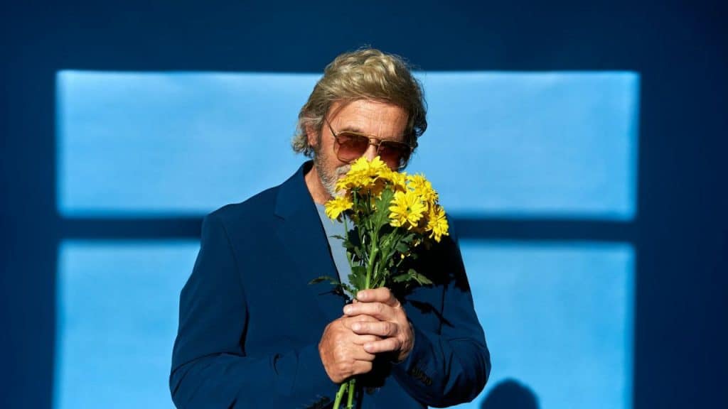 A man in a blue suit and sunglasses holds a bouquet of yellow flowers to his face against a blue wall.