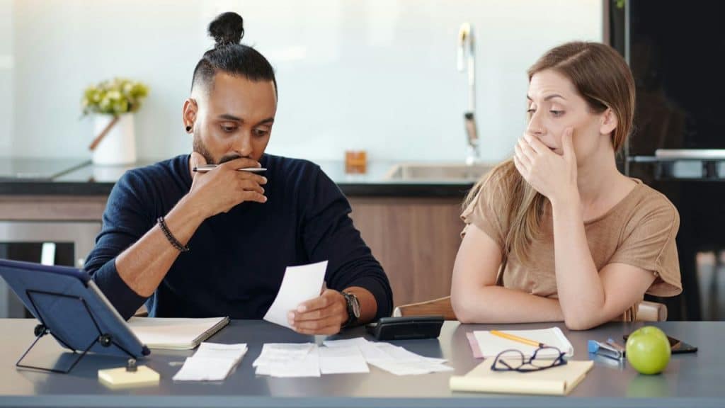 A man and a woman are sitting at a table with papers and a tablet, calculating finances, and the woman looks worried.