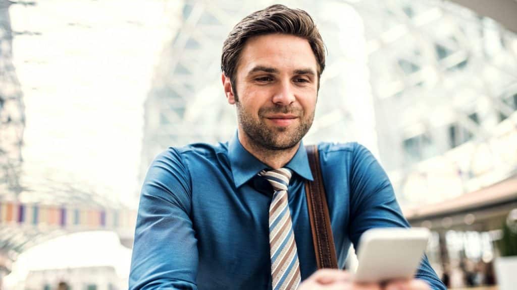 A well-dressed man smiles while looking down at his smartphone outdoors with modern buildings in the background.