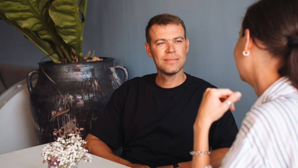 A man in a black shirt looks toward a woman sitting across a table.