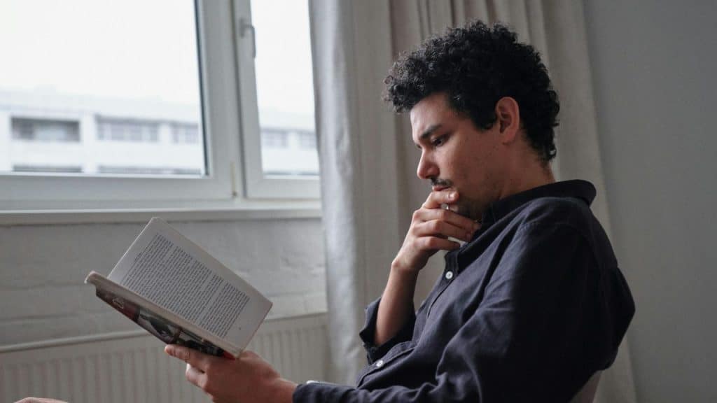 A serious man with curly hair and a mustache reads a book next to a window.