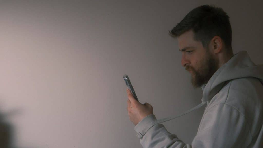 A bearded man in a light gray hoodie stands against a plain wall looking intently at his smartphone.