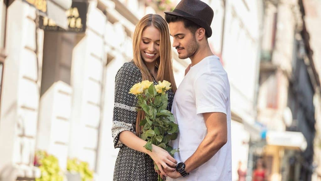 Smiling woman holding yellow roses next to a man in a hat outdoors.