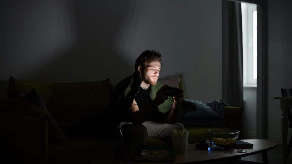 A man sitting on a couch at night looking at his phone with food on the table.