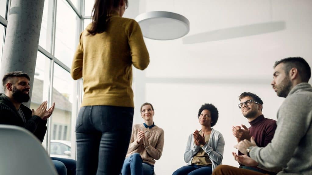 A group of diverse adults is sitting in a circle and clapping for a woman standing with her back to the camera.