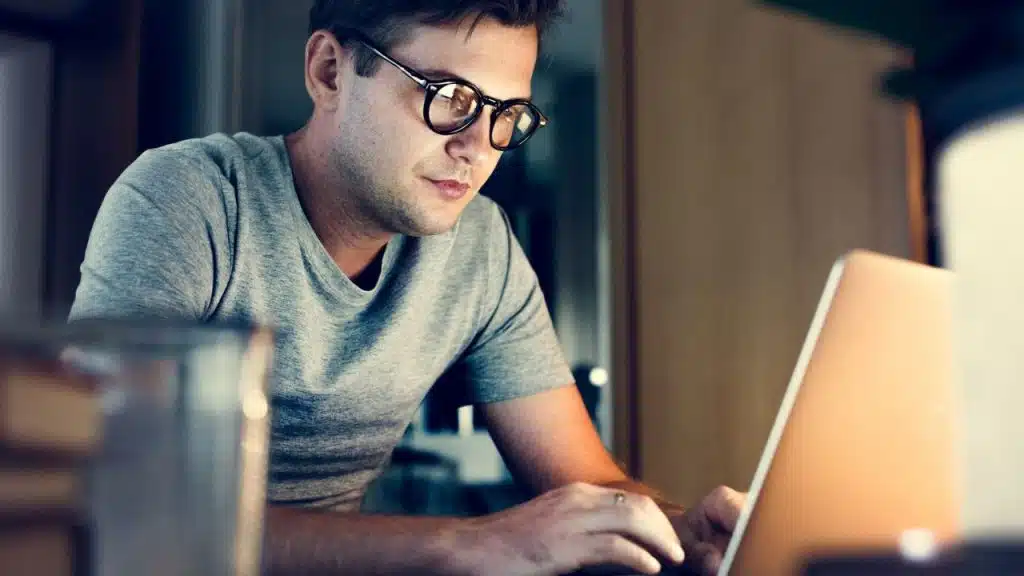 A man wearing round glasses is focused on working on a laptop in a dimly lit room.