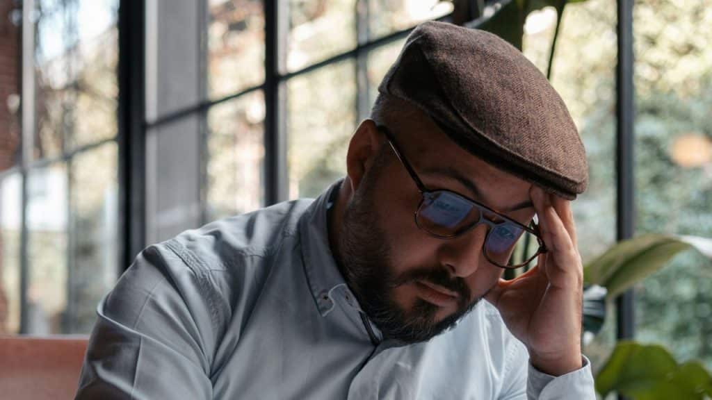 A bearded man in a white shirt and cap holds his head near a window.