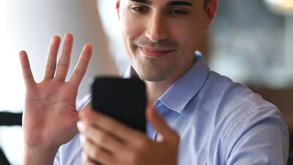 A man smiling and waving while having a video call on his phone.