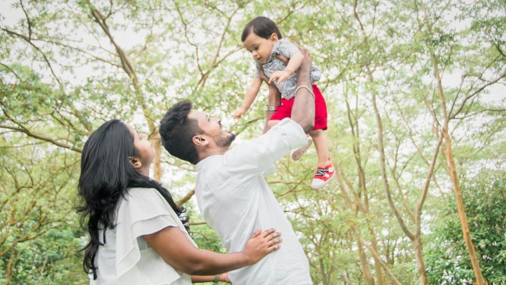 A smiling man lifts a young child wearing red shorts high into the air, while a woman watches nearby under a canopy of green trees.