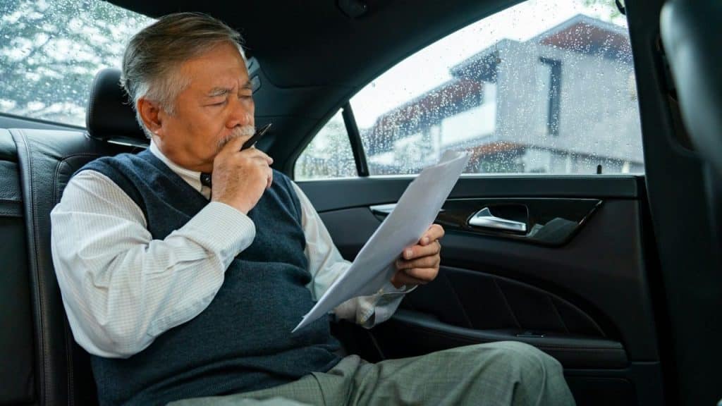 An older man in the back seat of a car examines a document while holding a pen to his mouth.