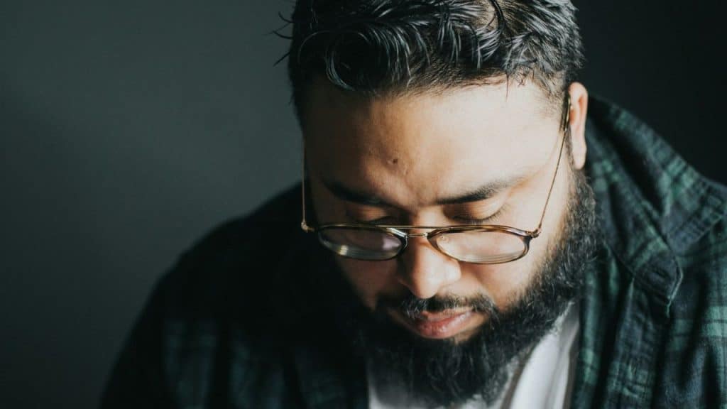 Close-up of a bearded man with glasses looking down in a dark setting.