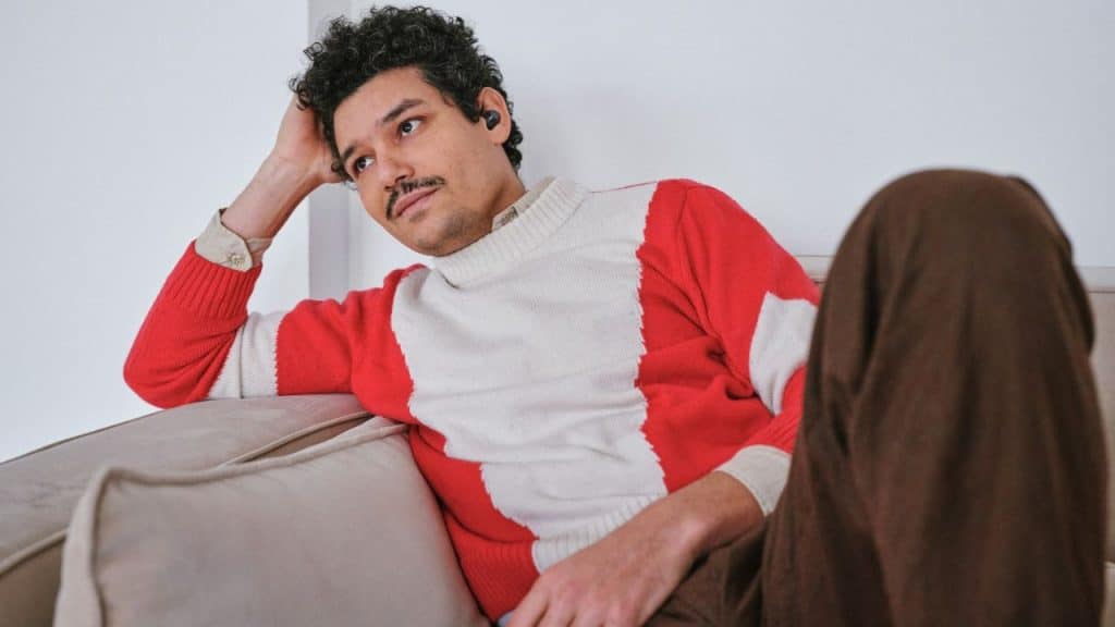 Man in a red and white sweater sitting on a couch, looking thoughtful or bored.