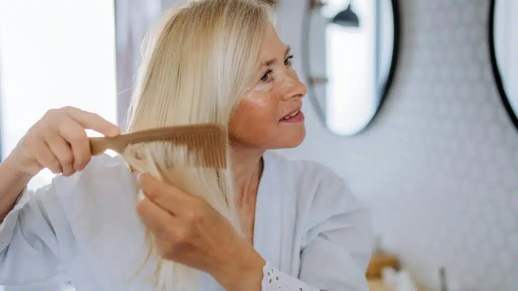A woman combing her long blonde hair in front of a mirror.