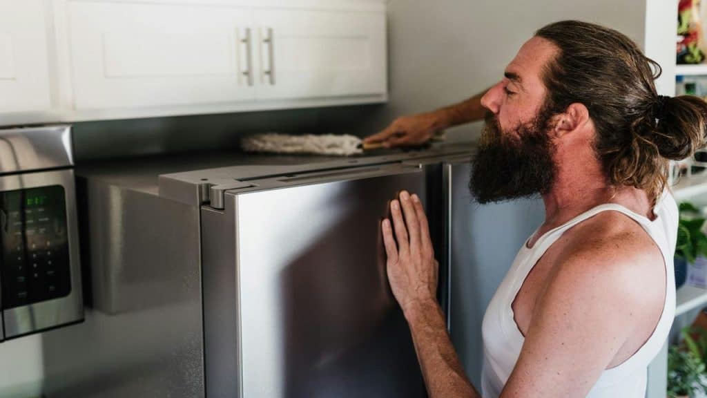 A man cleaning the top of a refrigerator in his kitchen.