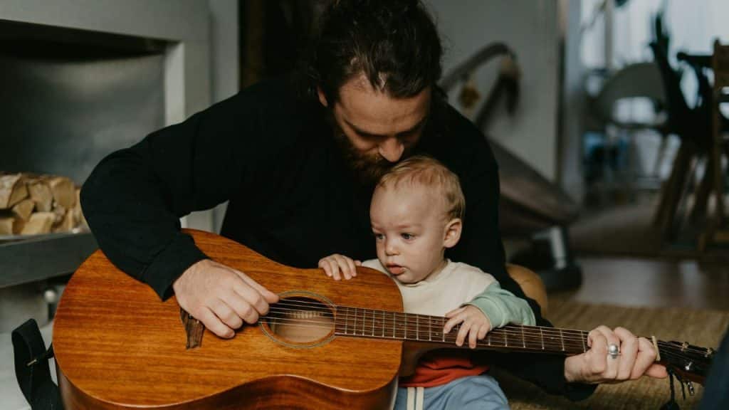 A man with long hair and a beard sits while holding a baby and a wooden acoustic guitar.