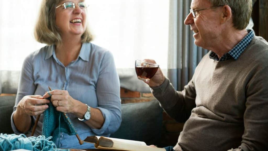 A man is reading while holding a glass of tea and smiles at a woman who is knitting, both sitting indoors.