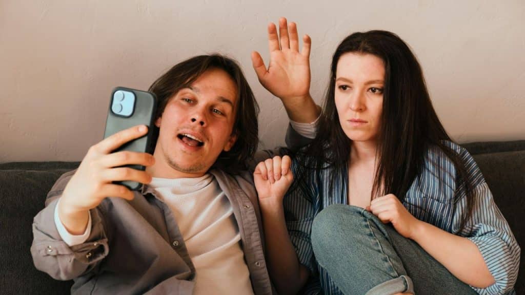 A man films himself and a woman on a phone; she waves with a bored expression.