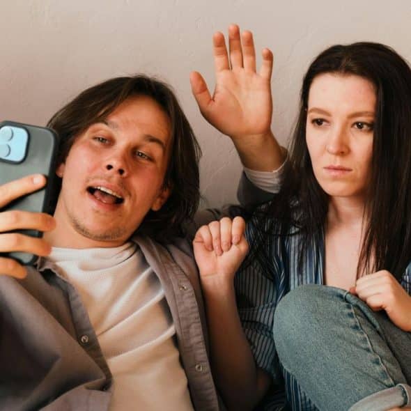 A man films himself and a woman on a phone; she waves with a bored expression.