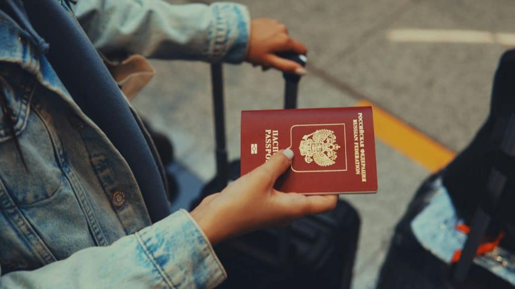 A close-up of two hands holding a deep red Russian passport over a dark piece of luggage and a denim jacket.