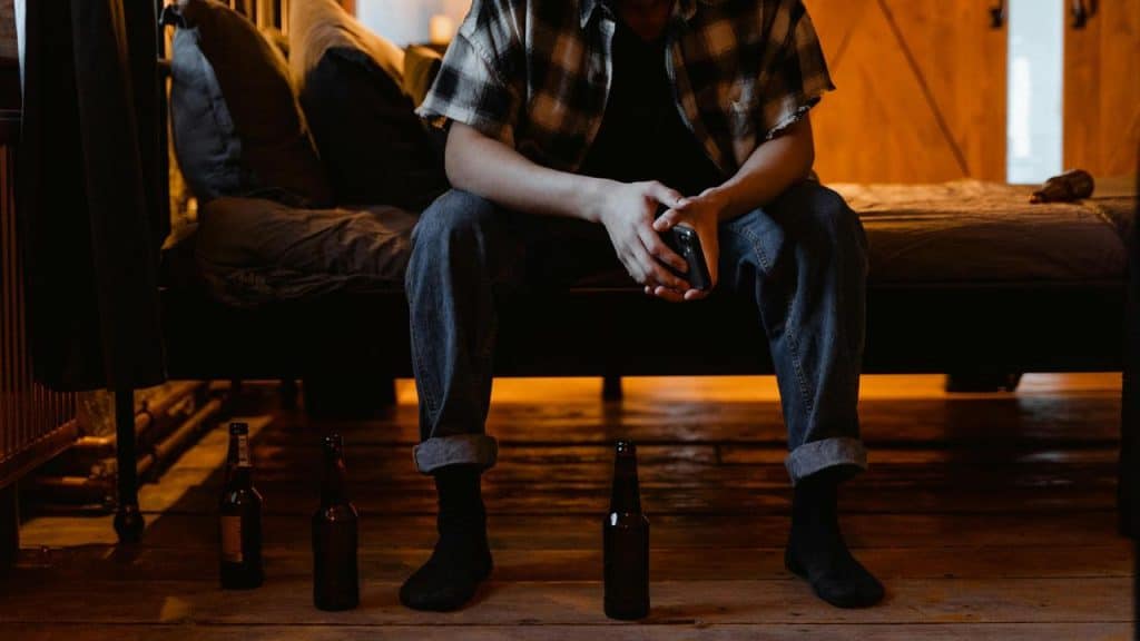 A person sitting on a bed holding a bottle with empty bottles on the floor.