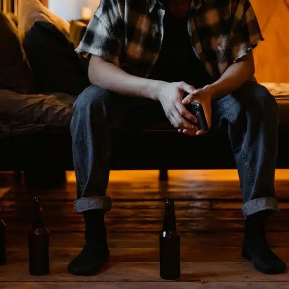 A person sitting on a bed holding a bottle with empty bottles on the floor.
