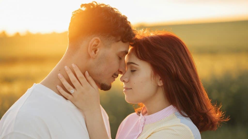 A close-up of a couple embracing and touching foreheads in a field, illuminated by a bright golden sunset.