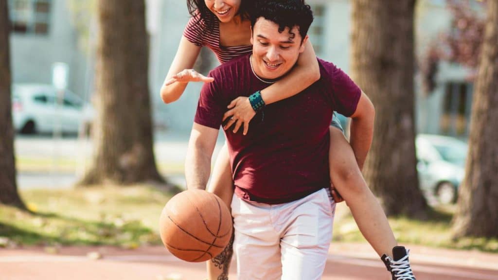 A playful couple outdoors, with one person piggybacking while holding a basketball.