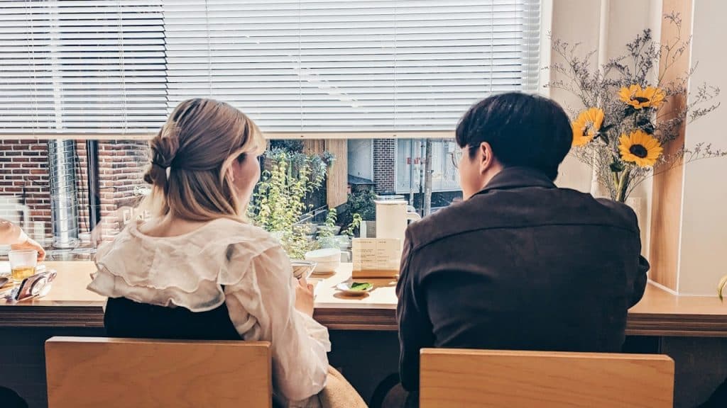 A blonde woman and a man with dark hair in a dark jacket are sitting facing a window with blinds at a coffee shop.