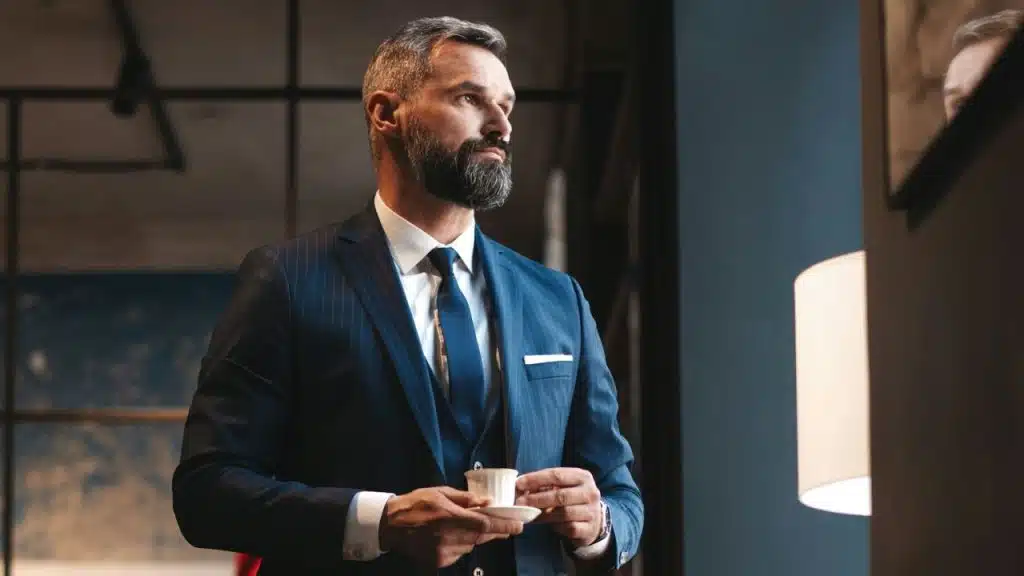 A well-dressed, bearded man in a blue pinstripe suit holds a small white teacup.