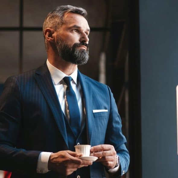 A well-dressed, bearded man in a blue pinstripe suit holds a small white teacup.