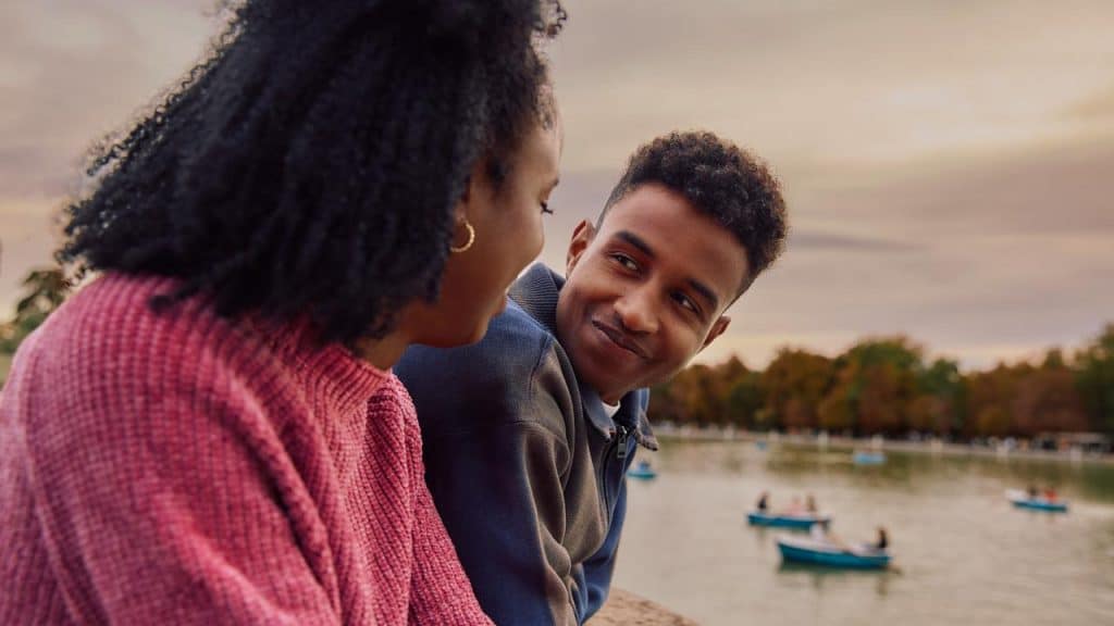 A couple sitting by a lake, smiling and looking at each other.