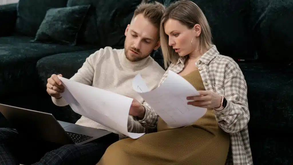 A couple sitting on the floor reviewing papers together near a laptop.