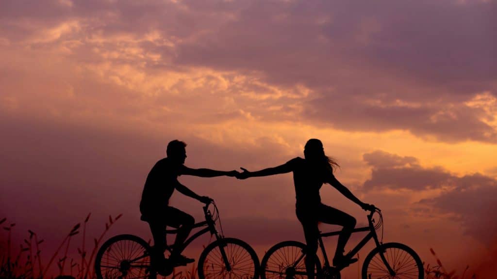 Silhouette of a couple riding bicycles and holding hands against a vibrant sunset sky.