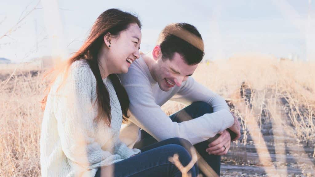 A joyful couple laughs together while sitting in a dry, grassy field under a bright sky.